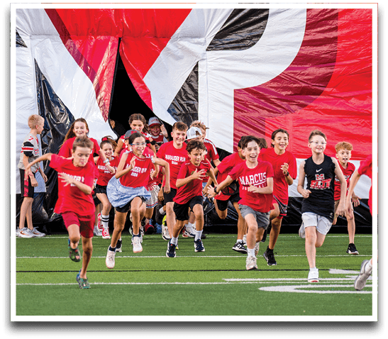Children wearing red and black sports shirts are running energetically out of a large inflatable tunnel onto a green field. Description generated by AI