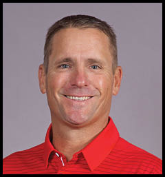 A person with short hair is smiling and wearing a red collared shirt with thin horizontal stripes against a plain gray background. Description generated by AI