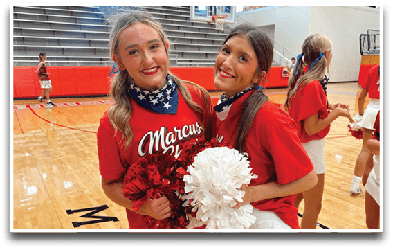 Two adults wearing red shirts and white skirts smile while holding red and white pom-poms in a gymnasium. Description generated by AI