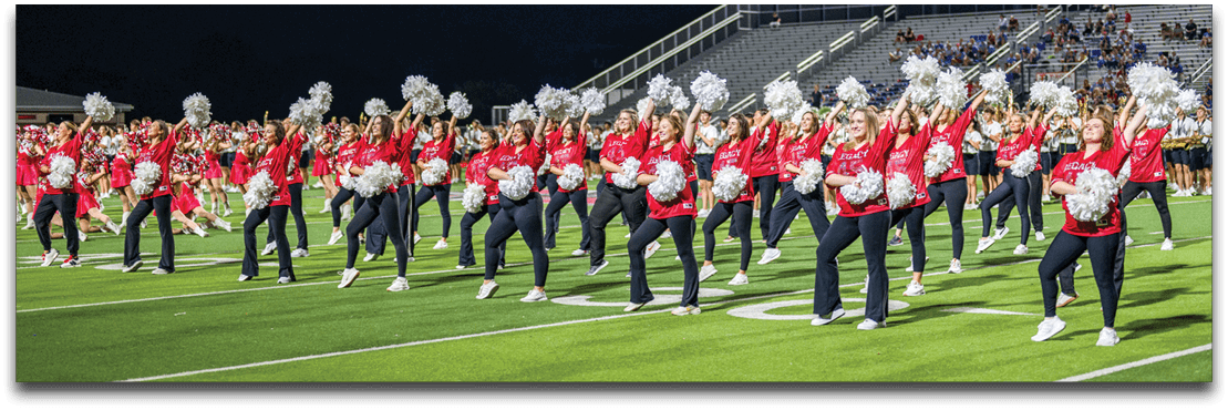 A large group of people dressed in red tops and dark pants are performing a synchronized cheerleading or dance routine on a well-lit sports field at night. Description generated by AI