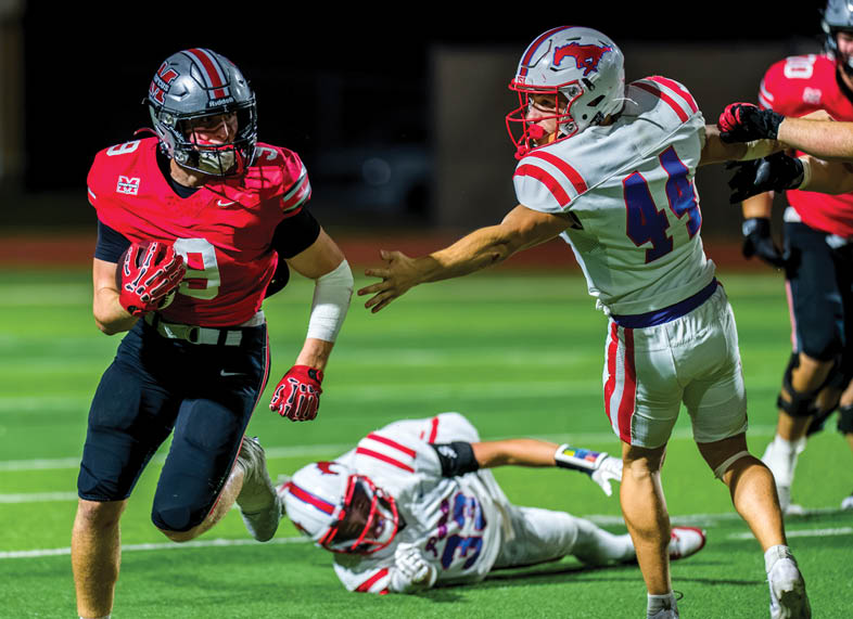 A person in a red and black football uniform is running with the ball while two people in white and red football uniforms attempt to tackle them on a green field. Description generated by AI