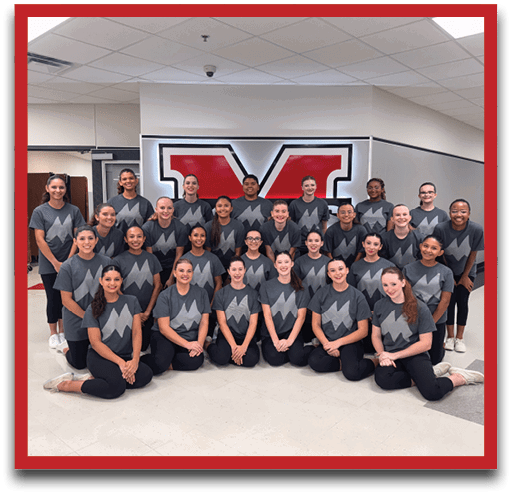 A group of smiling people wearing matching dark gray shirts with a stylized “M" logo pose together indoors in front of a wall with a large red and white graphic. Description generated by AI