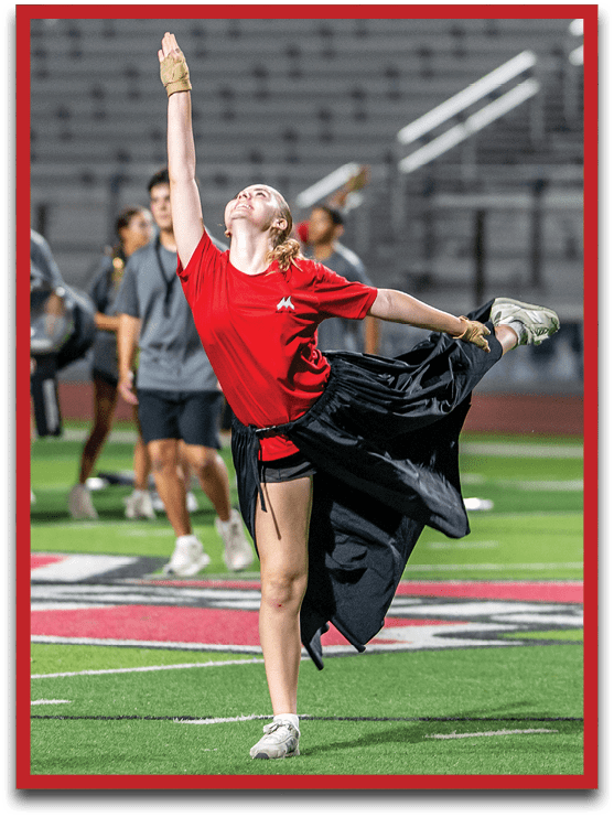 A person in a red shirt and black skirt is performing a dance or cheer routine on a sports field with a marching band standing in the background. Description generated by AI