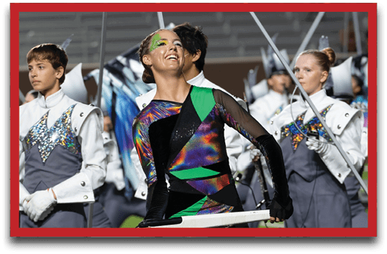 People in colorful and white uniforms are performing in a marching band or color guard event, with one person smiling and holding a flag pole. Description generated by AI