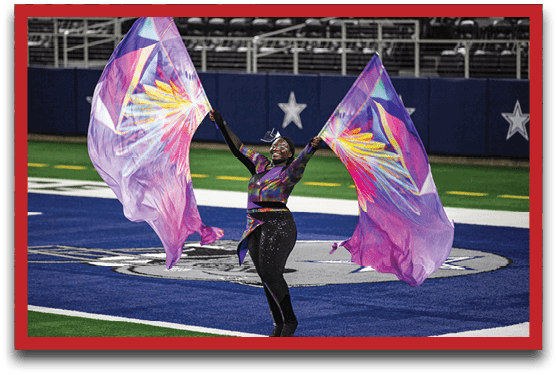 A person wearing a colorful outfit is performing on a football field while holding and waving two large purple flags with vibrant designs. Description generated by AI