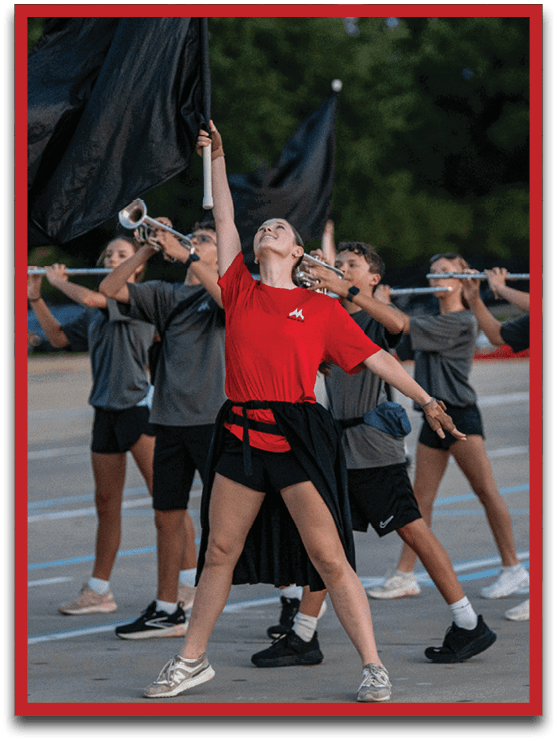 A group of people in casual athletic clothing are performing a coordinated marching band routine outdoors with one person in a red shirt prominently holding a flag. Description generated by AI