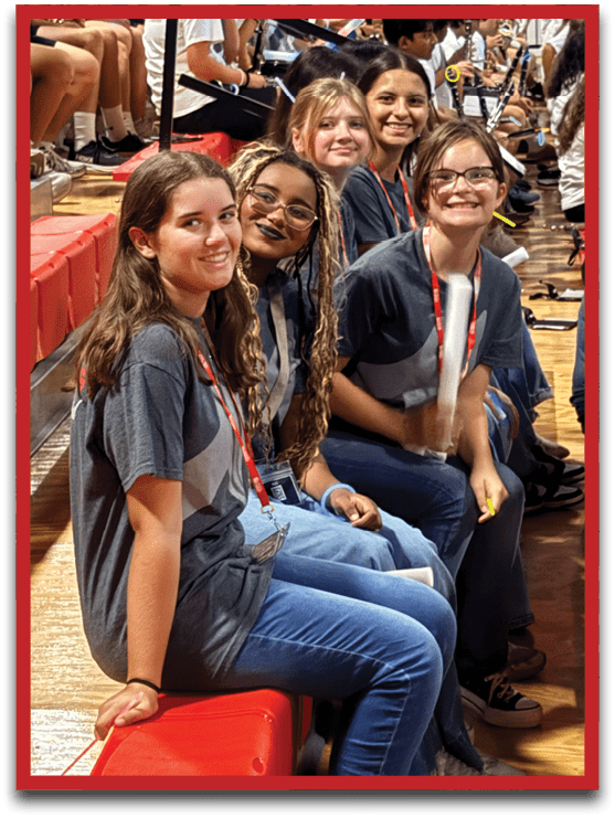 A group of people wearing casual clothing are sitting closely together on red bleachers in a gymnasium, some smiling and looking toward the camera. Description generated by AI
