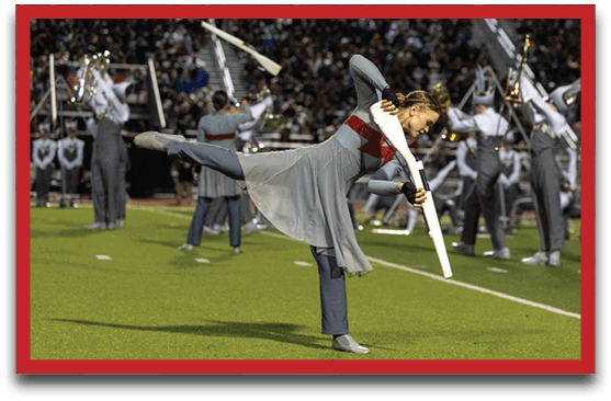 A person in a flowing gray and red costume is performing a balancing pose on one leg while holding a white rifle prop on a football field with a marching band in uniform playing instruments in the background. Description generated by AI