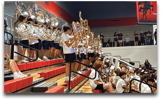 A group of people in matching white shirts and dark shorts are playing brass instruments on bleachers in a gymnasium while an audience watches from nearby seats and an upper balcony. Description generated by AI