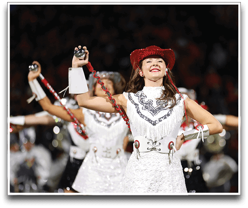 A person wearing a red sequined hat and a white fringed outfit is smiling and holding a baton with a red spiral handle, likely performing in a marching band or dance routine. Description generated by AI