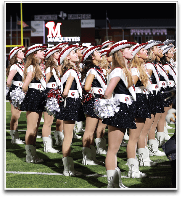 A group of adults dressed in coordinated sequined outfits, white boots, and hats are standing in formation on a football field with pom-poms, likely performing a dance or drill routine. Description generated by AI