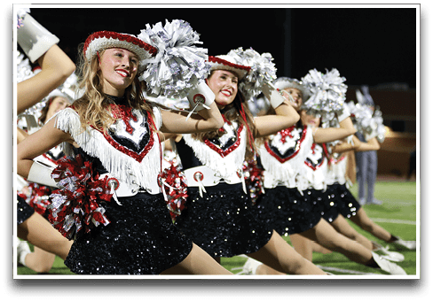A line of adults dressed in sequined skirts, fringed tops, and decorative hats perform a synchronized dance routine on a field while holding pom-poms. Description generated by AI
