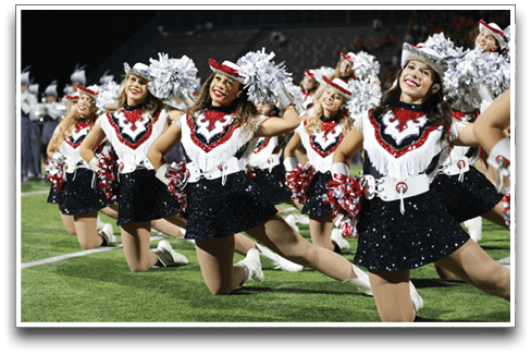 A group of people dressed in sequined skirts, fringed tops, and hats are kneeling on a football field holding pom-poms while a marching band plays in the background. Description generated by AI