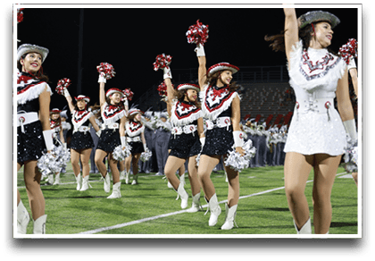 A group of adults dressed in sequined dance uniforms and hats perform a synchronized routine on a football field at night with a marching band in the background. Description generated by AI