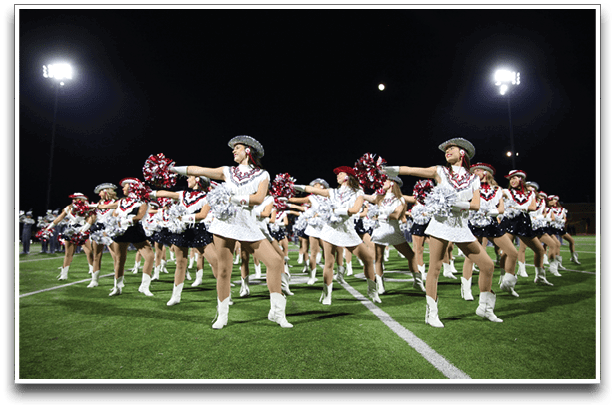 A large group of people dressed in coordinated white and black outfits with hats and boots are performing a synchronized dance routine on a lit outdoor field at night. Description generated by AI