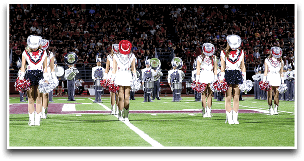 A group of people dressed in coordinated outfits with hats and pom-poms are performing a synchronized routine on a football field with a marching band in the background. Description generated by AI