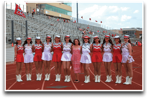 A group of people dressed in coordinated red and white dance or cheer uniforms with hats and boots stand on a running track in front of bleachers, with one person in a pink dress standing in the middle. Description generated by AI
