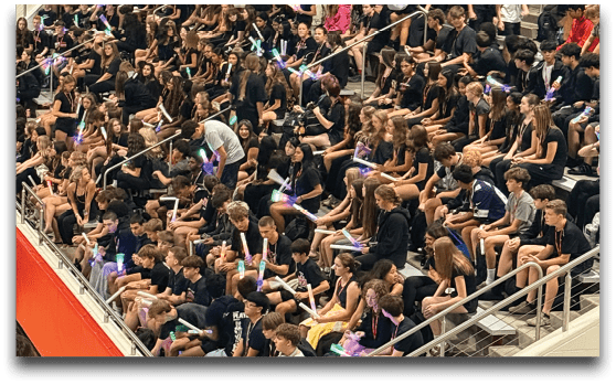 A large crowd of people wearing mostly dark clothing are seated closely together in bleachers inside a gymnasium, many holding glowing light sticks. Description generated by AI
