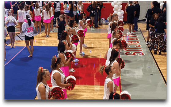 A group of people dressed in white tops and pink skirts are lined up on a gymnasium floor holding red pom-poms, appearing to perform a cheerleading routine. Description generated by AI