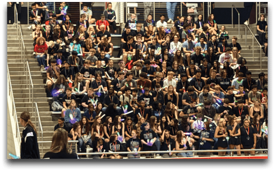 A large group of people wearing matching dark outfits is seated closely together on bleachers inside a gymnasium with an American flag visible on the right side. Description generated by AI