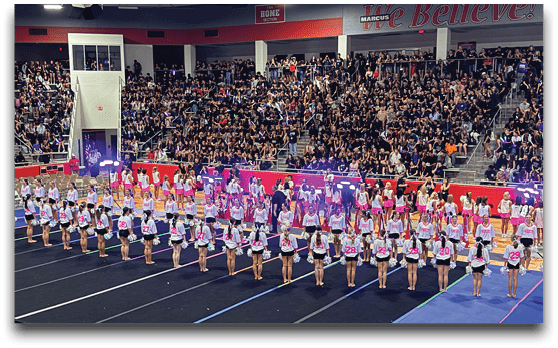 A large group of people in coordinated outfits are standing in organized rows on a gymnasium floor with a packed audience watching from the bleachers. Description generated by AI