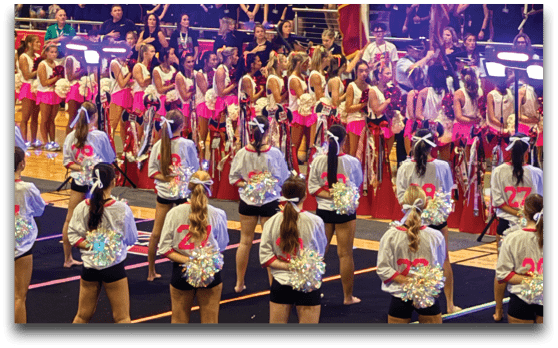 A large crowd of people wearing black shirts stands in bleachers while a group of people in pink and white cheerleading outfits perform or stand in front of them in a gymnasium setting. Description generated by AI
