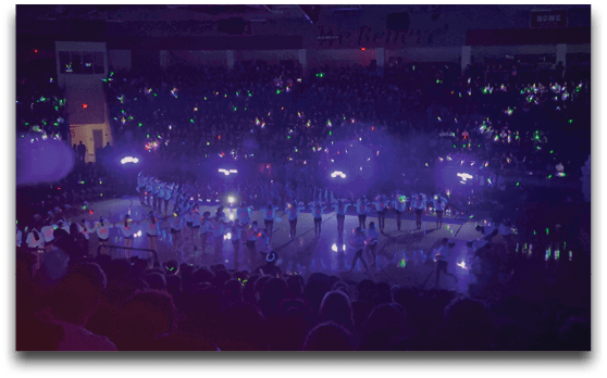A large group of people in white outfits perform a coordinated dance or routine on a brightly lit stage in front of a dark audience holding glowing green and blue lights. Description generated by AI