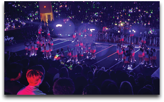 A large crowd watches a cheerleading performance in a dimly lit indoor arena with performers wearing bright outfits and holding glowing sticks. Description generated by AI