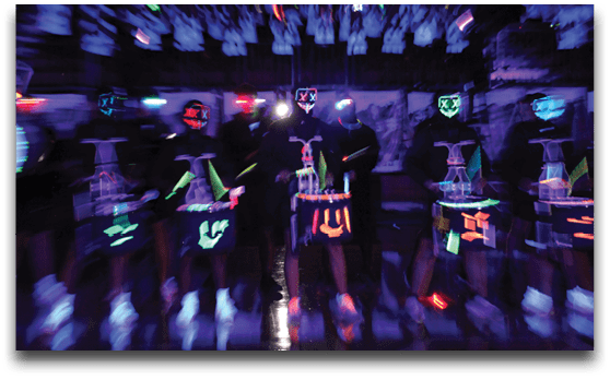 People wearing dark clothing with glowing neon paint and masks stand in a dimly lit room with blurred motion effects. Description generated by AI