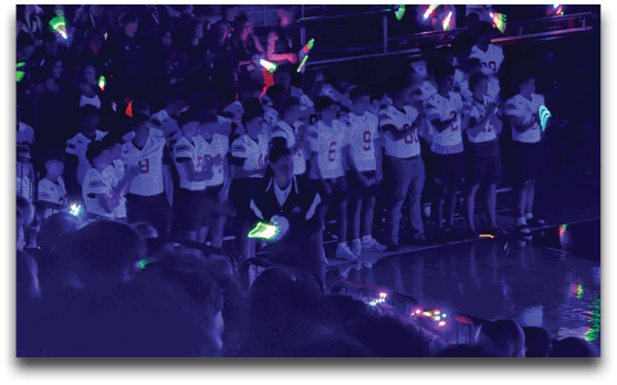 A group of people wearing matching sports jerseys stand in a row inside a dimly lit gymnasium with colorful glowing sticks and lights around them. Description generated by AI