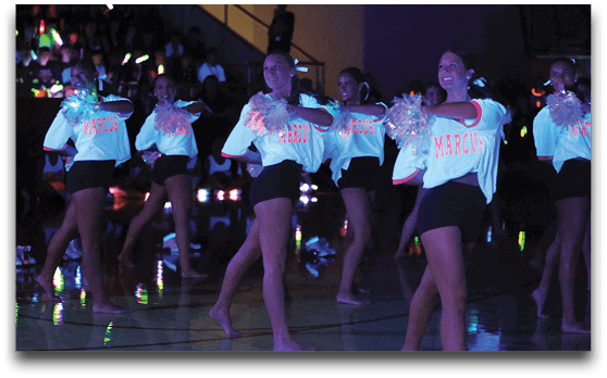 People in matching shirts and shorts perform a cheerleading routine with pom-poms on a gym floor illuminated by colorful lights and glow sticks in the background. Description generated by AI
