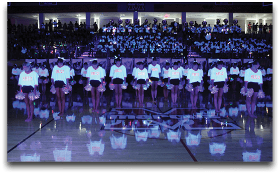 A group of people in matching light-up shirts and holding pom-poms stand in a line on a gymnasium floor with a crowd visible in the background. Description generated by AI