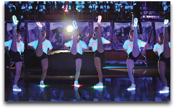 People in matching outfits perform a high kick routine on a shiny floor with their shoes glowing under blacklight in a gymnasium setting. Description generated by AI
