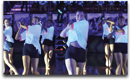 A group of adults wearing white shirts and black shorts are performing a synchronized dance routine on a dimly lit stage. Description generated by AI