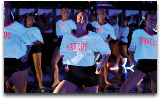 A group of people wearing white shirts with “MARCUS" printed in red and black shorts are performing a synchronized dance routine under dim lighting. Description generated by AI