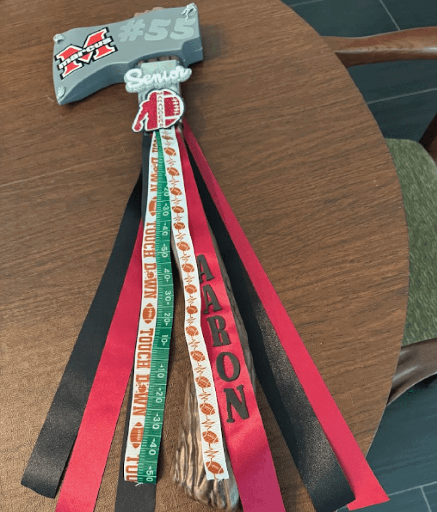 A decorated senior keychain with red and black ribbons, a name spelled “AARON" on one ribbon, and football-themed ribbons on a wooden table. Description generated by AI