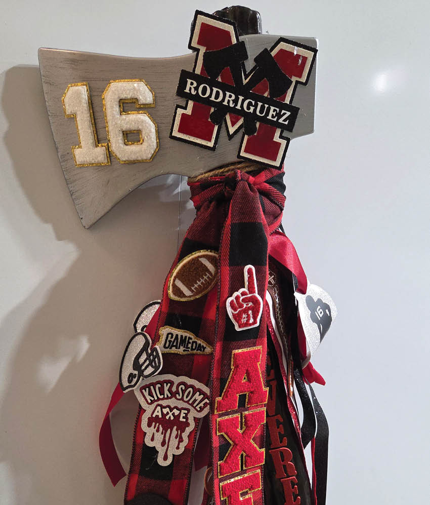 A decorative axe head painted silver with the number 16 and a large red and black letter M with the name Rodriguez, adorned with red and black ribbons and patches including a football, foam finger, and phrases like “GAMEDAY" and "KICK SOME AXE." Description generated by AI