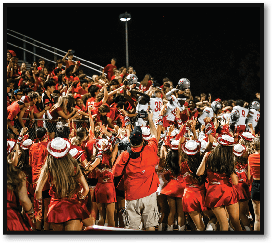 A large crowd of people wearing red clothing and hats enthusiastically cheer and raise their hands toward a group of football players in white uniforms standing on the field at night. Description generated by AI
