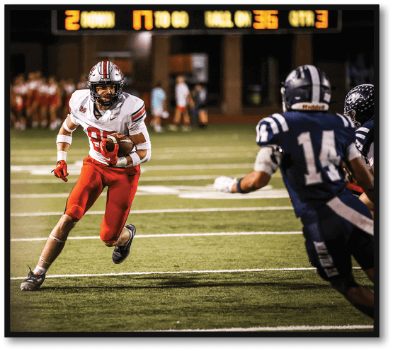 A person in a white and red football uniform is running on a field while holding a football, with a blurred group of people in the background. Description generated by AI