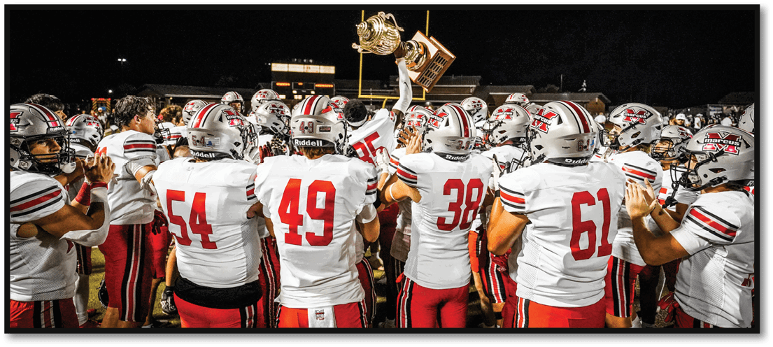 A group of people wearing white and red football uniforms are gathered closely together at night, with one person holding up a trophy. Description generated by AI
