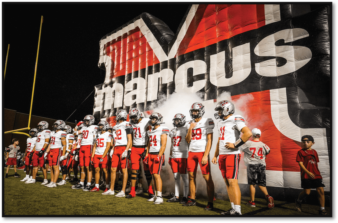 A group of people in matching sports uniforms stand in a line on a field near a large inflatable tunnel with smoke coming out. Description generated by AI