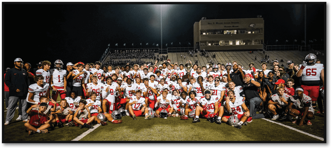 A large group of people wearing football uniforms and casual clothing pose together on a football field at night under bright stadium lights. Description generated by AI