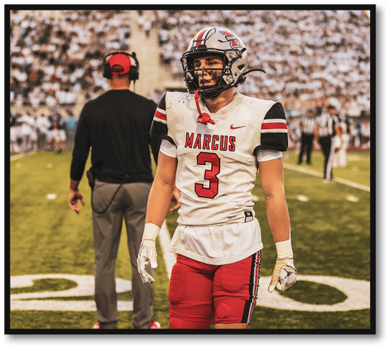 A person in a football uniform with the name Marcus and number 3 stands on a field while another person wearing a headset and black shirt faces away in the background with a crowd in the stands. Description generated by AI