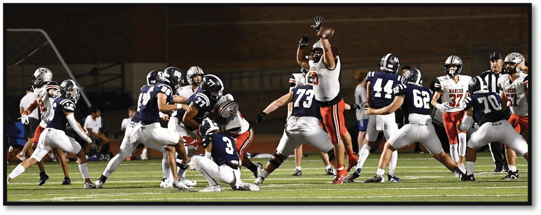 People in football uniforms are actively playing on a field under stadium lights with one player in white and red jumping to catch a football while others in blue and white attempt to block or tackle. Description generated by AI