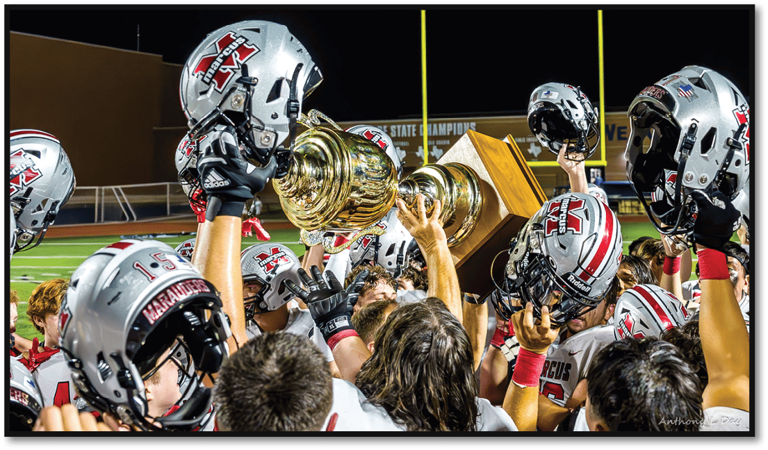 A group of people wearing white and red football uniforms are gathered closely together at night, with one person holding up a trophy. Description generated by AI