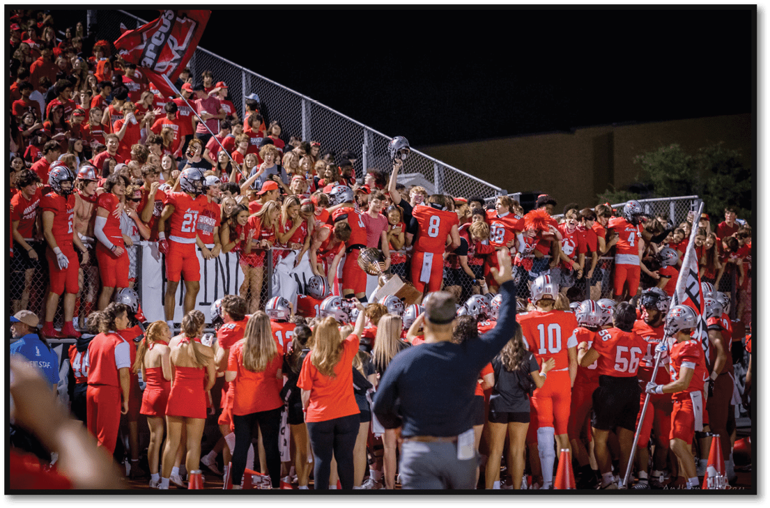 A group of athletes in red uniforms and a crowd of people wearing red gather excitedly near a chain-link fence at a nighttime sports event. Description generated by AI