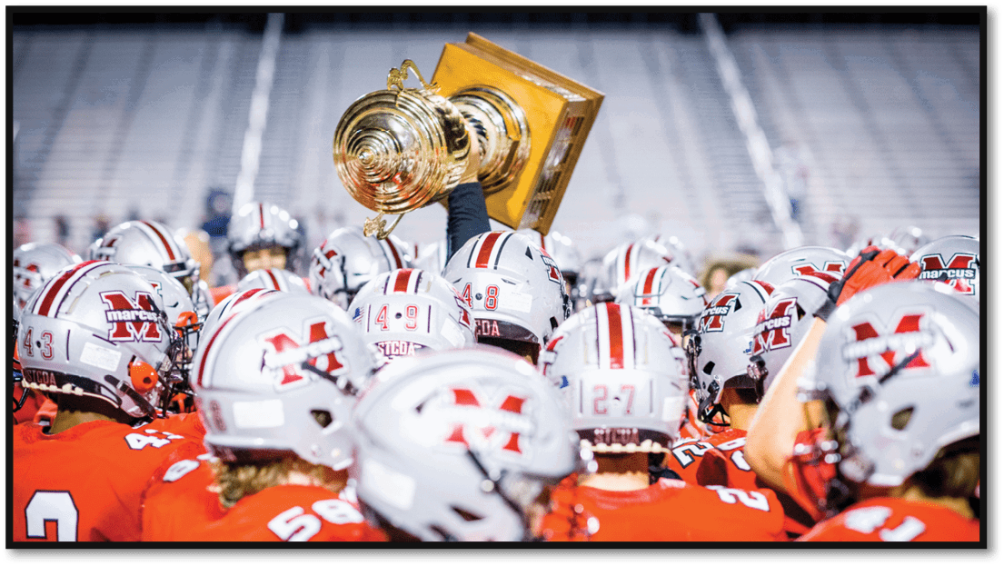 A group of people wearing red sports uniforms and helmets are gathered closely together with one person holding up a large trophy in a stadium. Description generated by AI