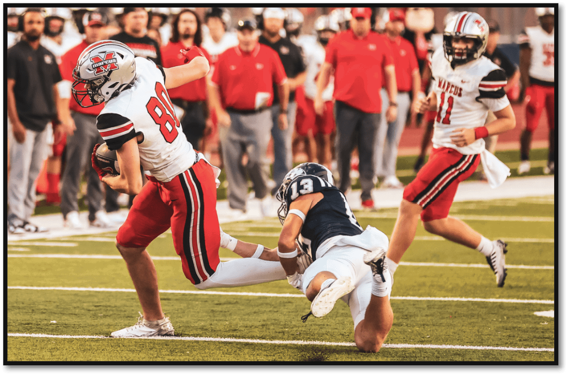 A person in a white and red football uniform is being tackled by another person in a navy blue and white uniform on a football field with a crowd and team members in the background. Description generated by AI