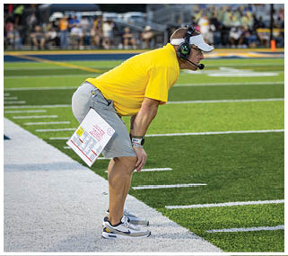 A man wearing a yellow shirt and a headset is bending over on a football field. AI generated content