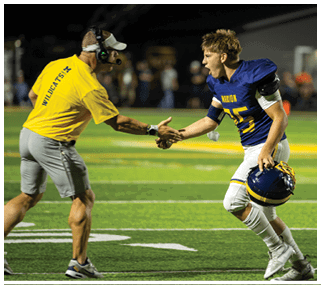Two men on a football field, one wearing a blue jersey and the other wearing a yellow shirt. They are shaking hands and one of them is wearing a helmet. AI generated content